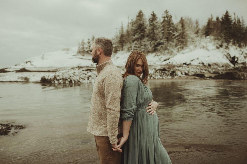 A couple stands by the water's edge with snow-covered hills and pine trees in the distance.