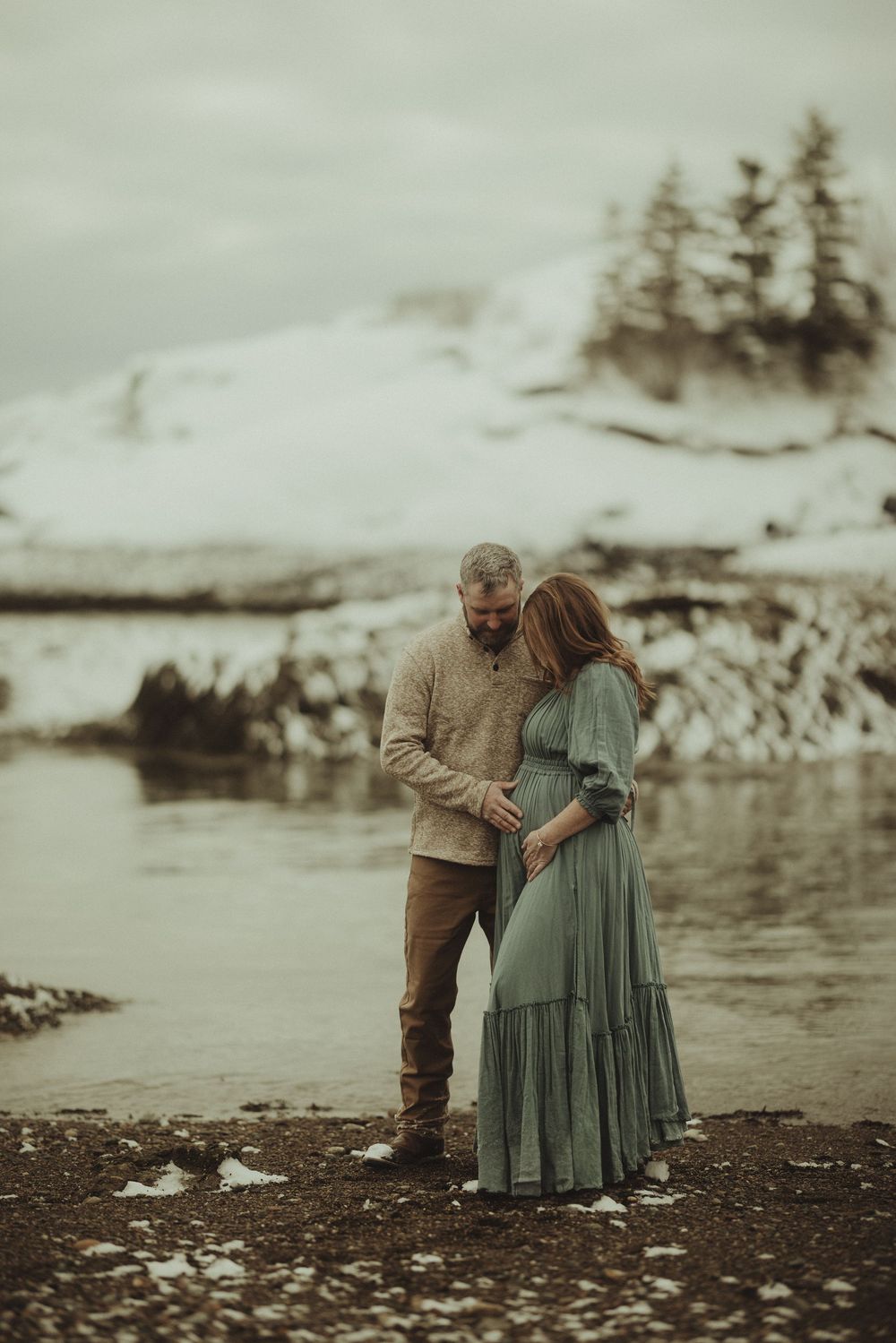 A couple shares an intimate moment on a rocky beach with snow-capped evergreens in the background.