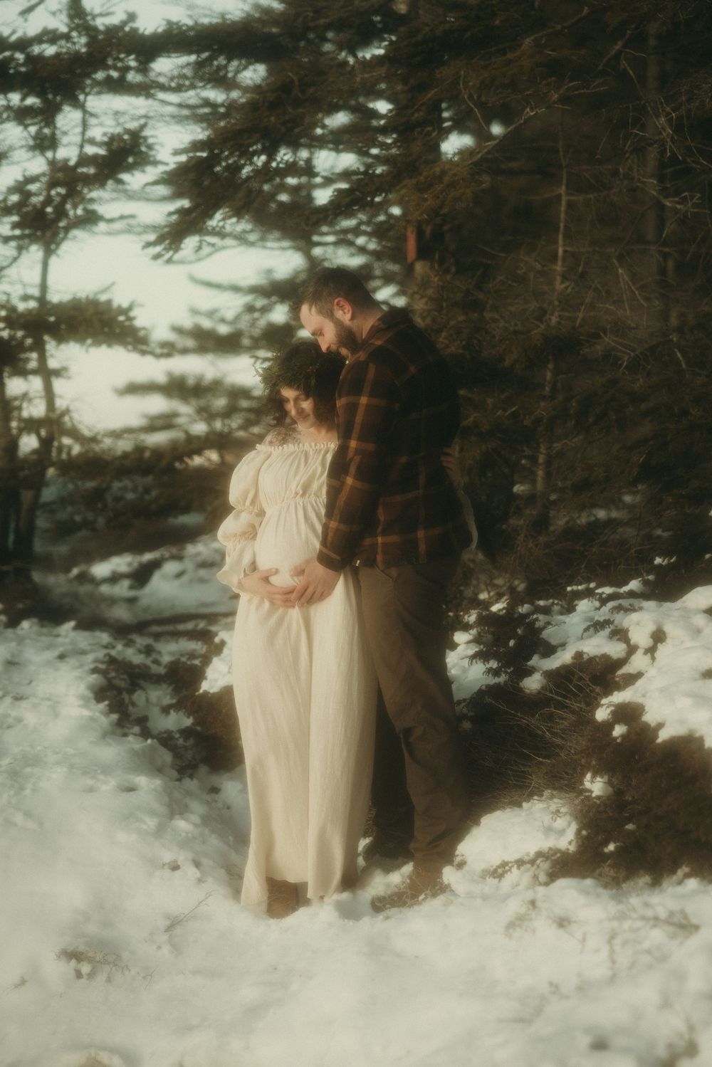 A romantic winter photoshoot with a couple standing in snowy woods near a cabin.