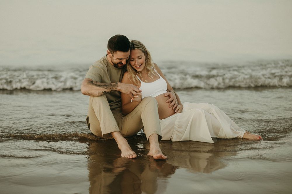 A romantic beach photoshoot features a couple sitting in gentle ocean waves.