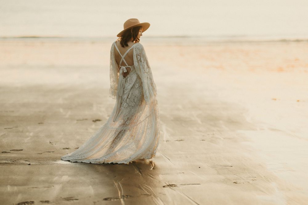Dreamy beach maternity portrait with flowing white dress and straw hat against a soft sunset backdrop.