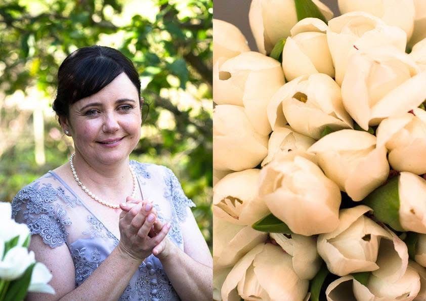 Smiling bride in lace dress holds hands beside a bouquet of white tulips outdoors.