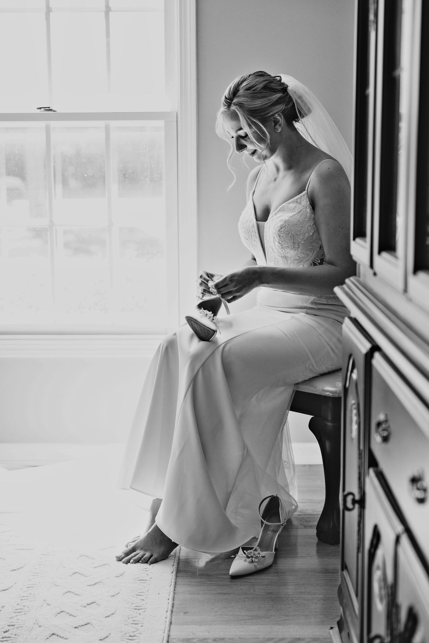 Black and white photo of bride sitting barefoot by window in wedding dress before ceremony.