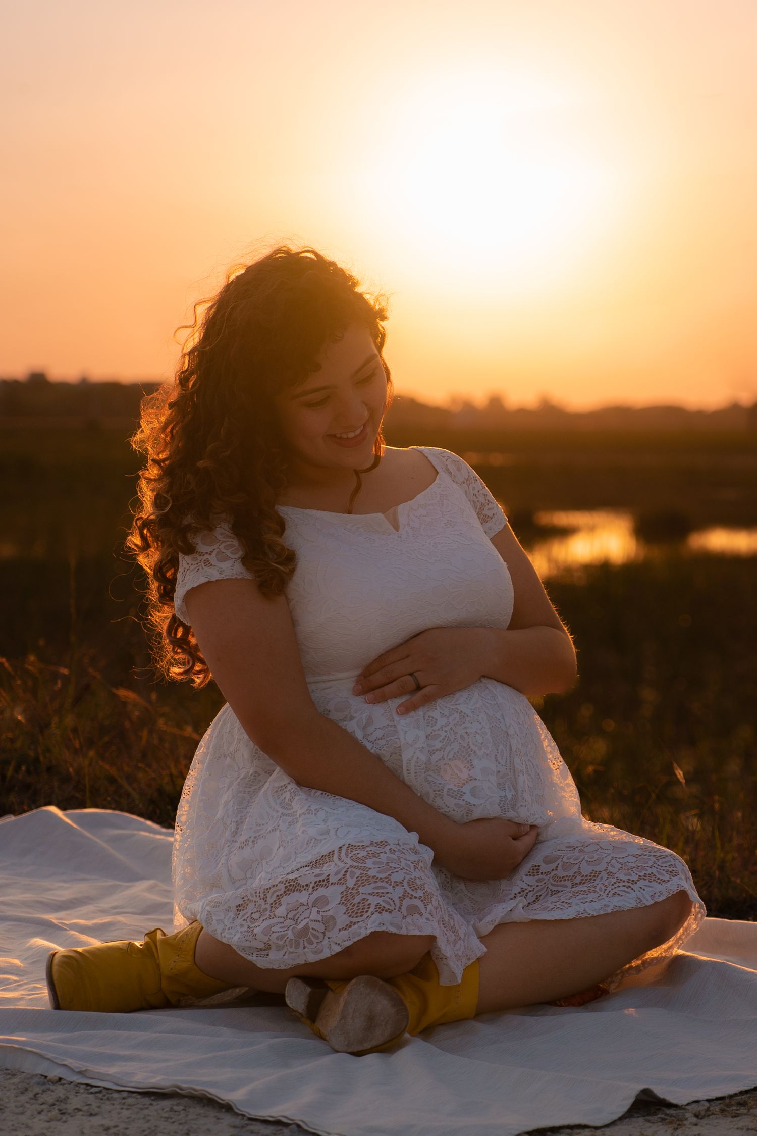 Pregnant woman in white lace dress sitting outdoors during golden sunset holding her baby bump.