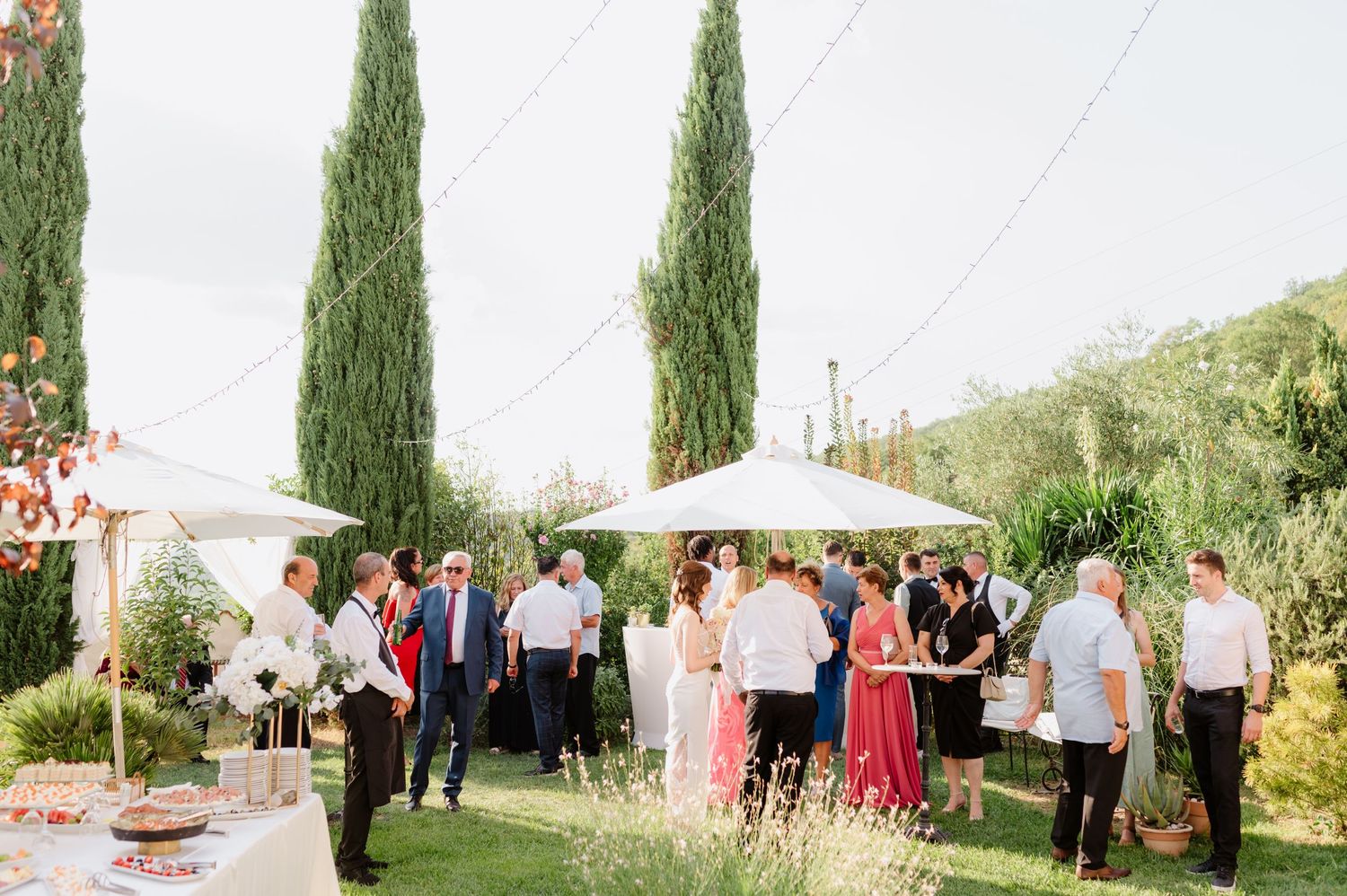 Garden wedding reception with guests mingling under umbrellas and cypress trees.