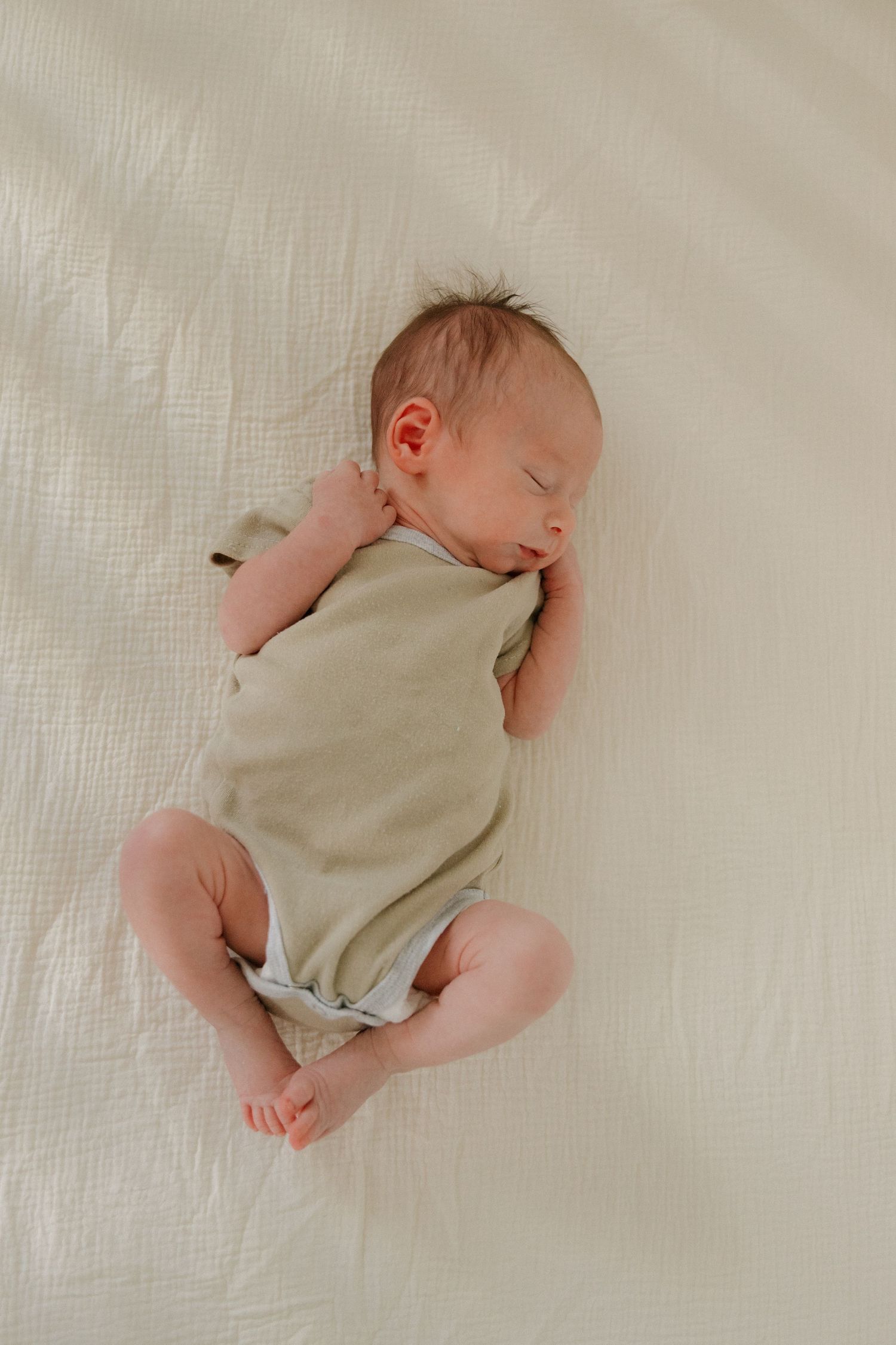 A sleeping newborn baby wearing a beige onesie lies peacefully on white textured bedding.