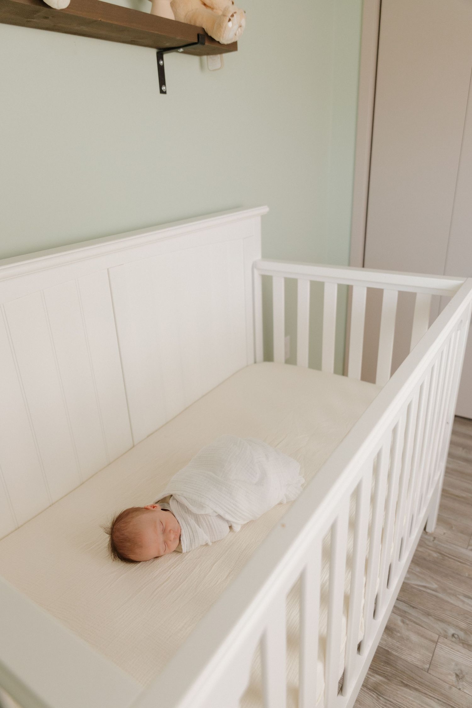 A sleeping baby in white clothing rests peacefully in a white wooden crib against a wall with floating shelf.