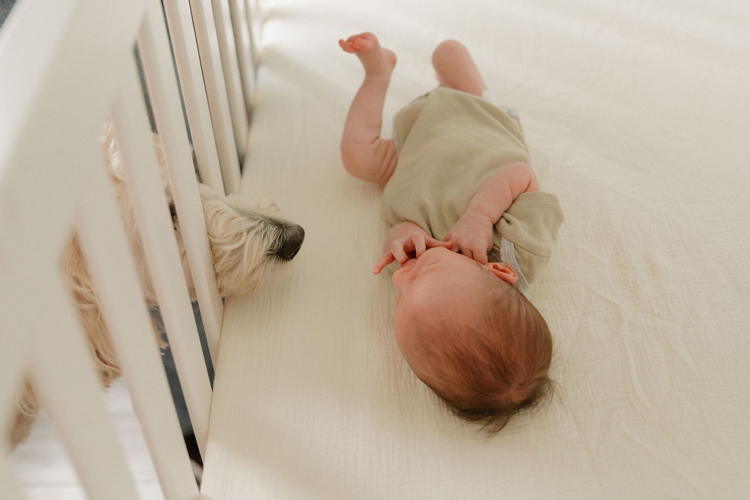 Baby in light green onesie lying on white nursery bedding near white crib rails.