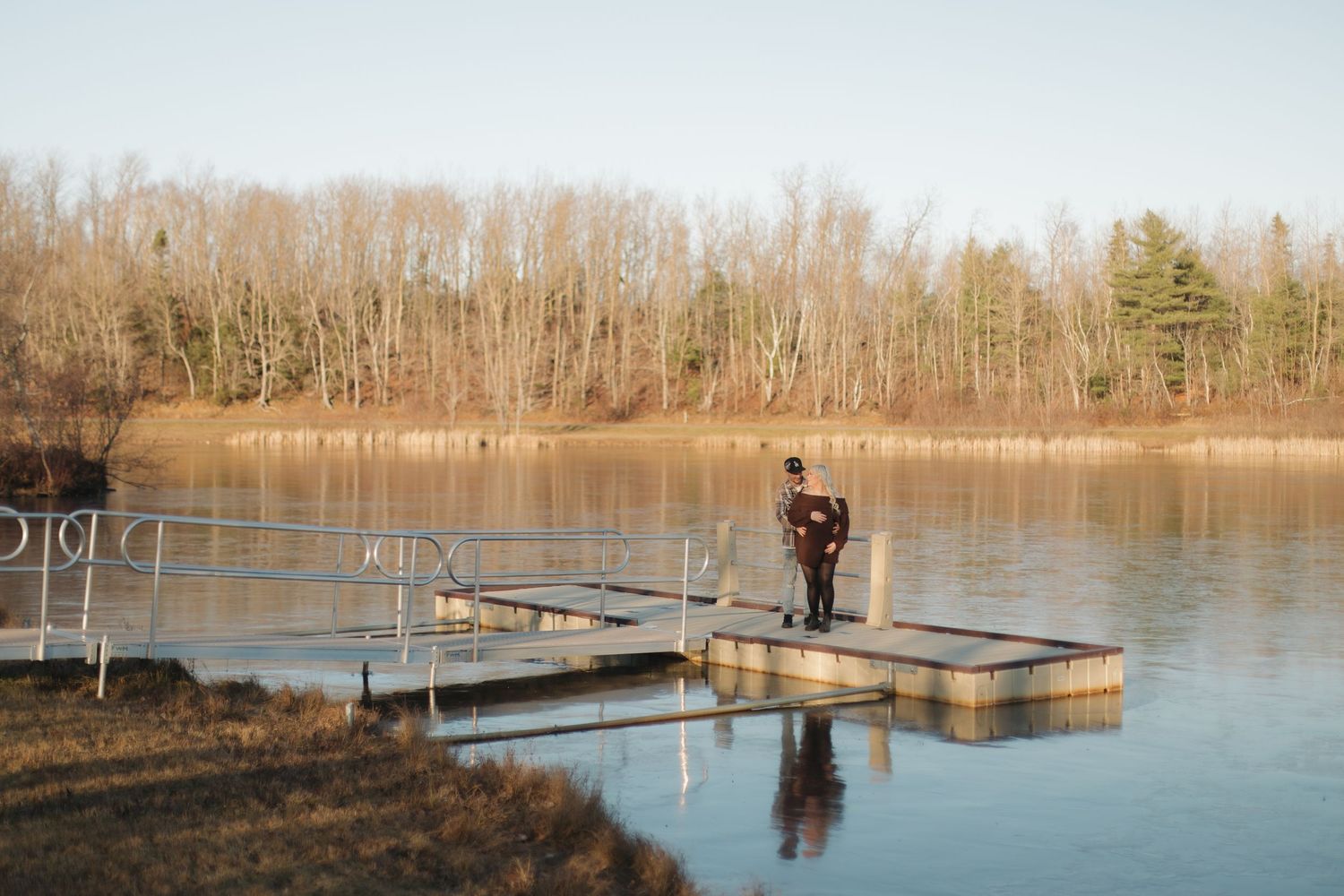 Two people stand close together on a wooden dock overlooking a serene lake surrounded by bare winter trees.