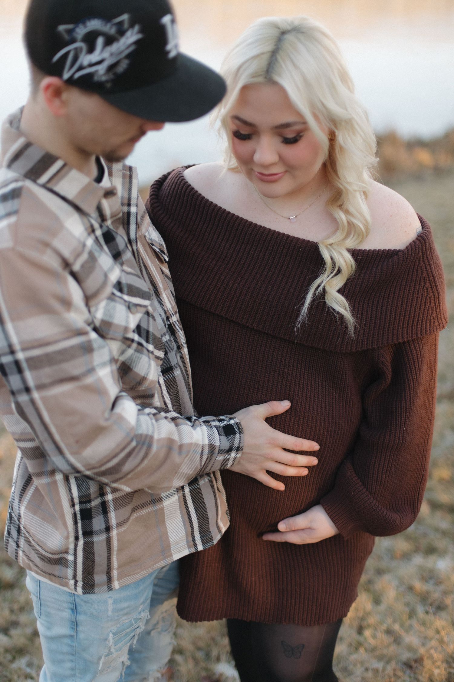 A couple shares an intimate moment outdoors as they lovingly touch a baby bump in a brown sweater maternity photo.
