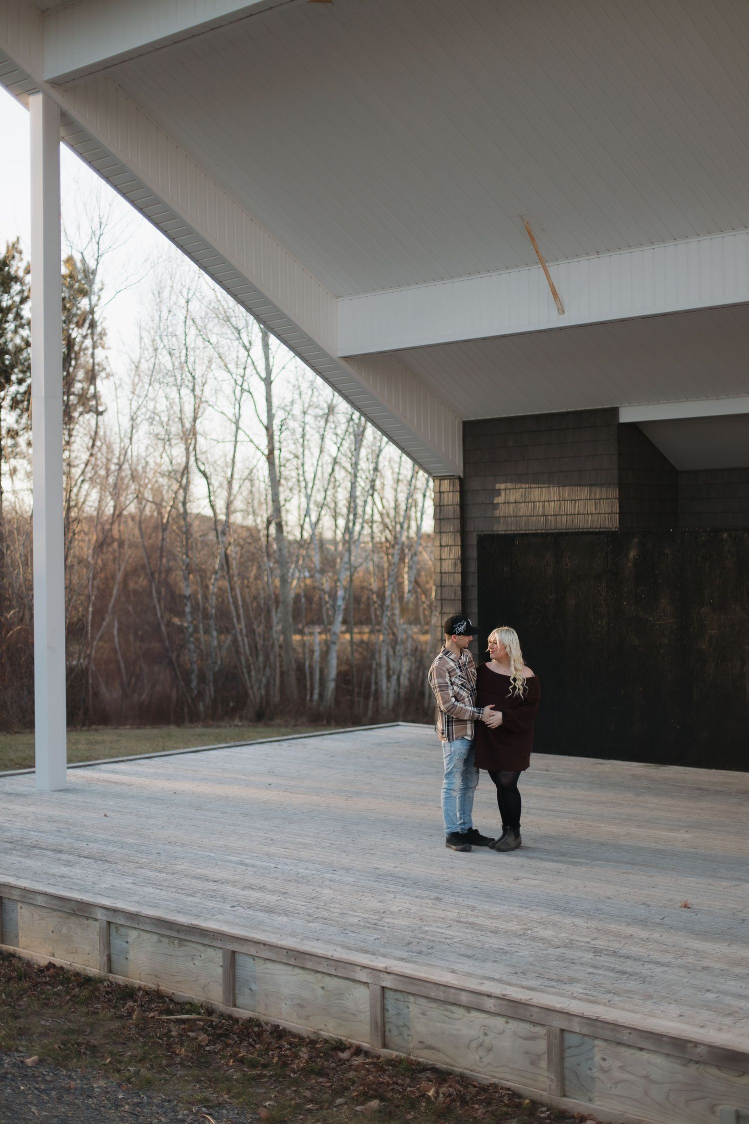 A couple stands on a modern covered patio deck with white columns and dark stone wall during winter sunset.