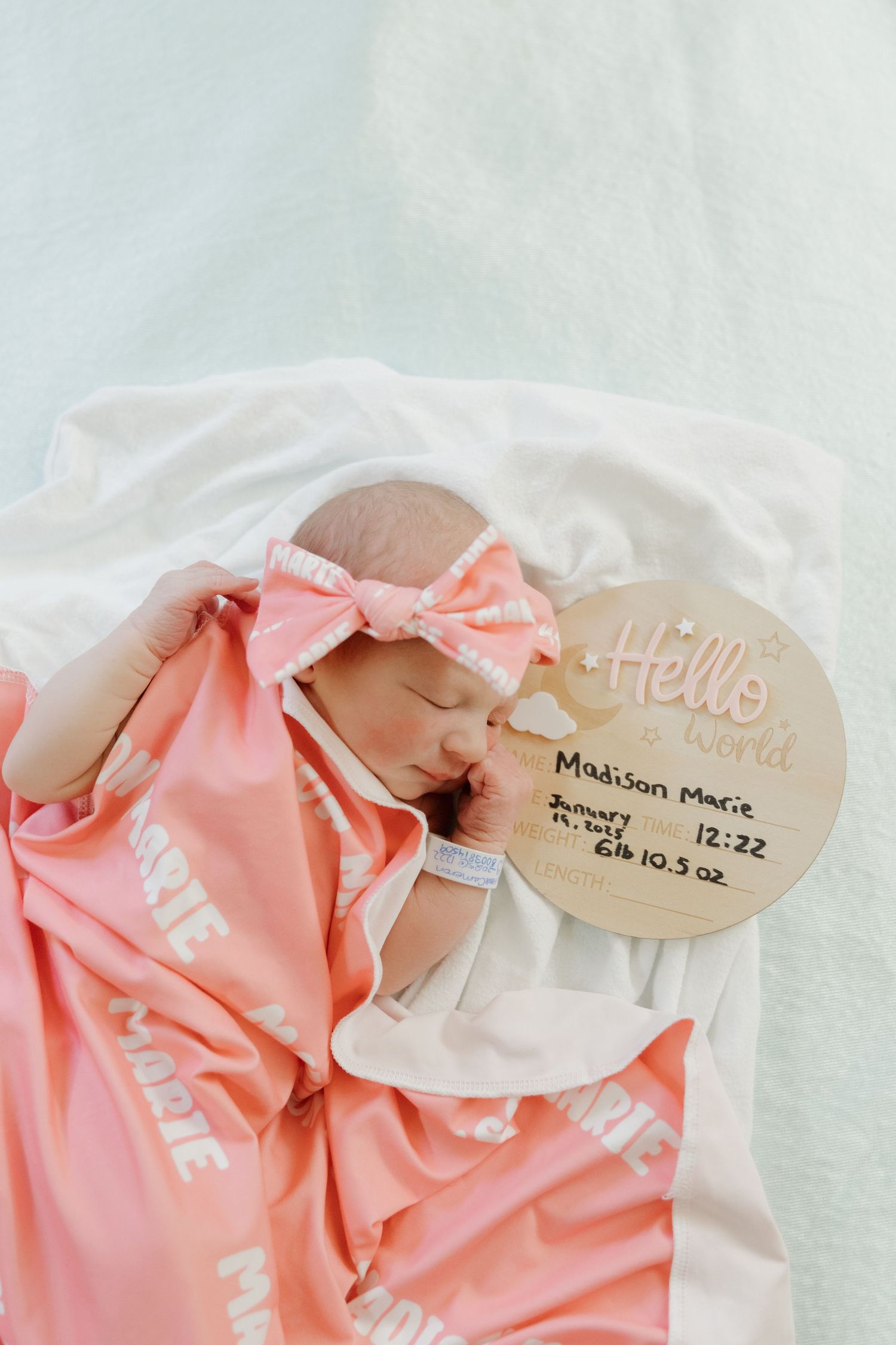 A newborn baby wrapped in coral pink blanket with matching bow headband next to a wooden name plate.