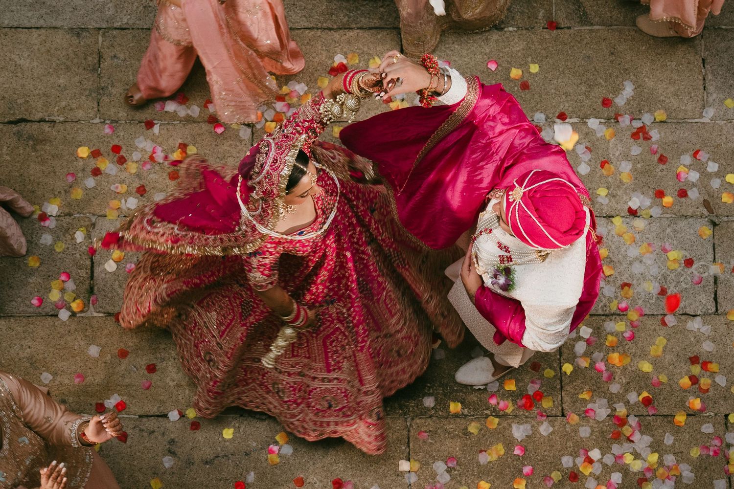 Aerial view of Indian bride and groom in vibrant red wedding attire surrounded by colorful confetti on stone steps.
