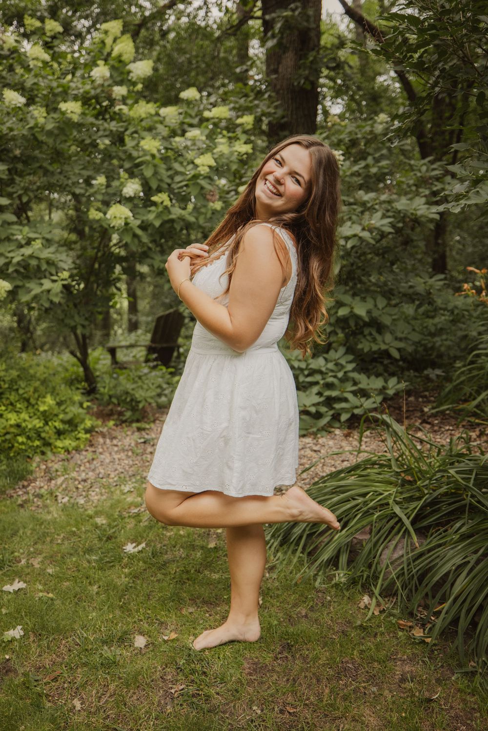 Smiling young woman in white sundress standing barefoot on one leg in a lush green garden setting.