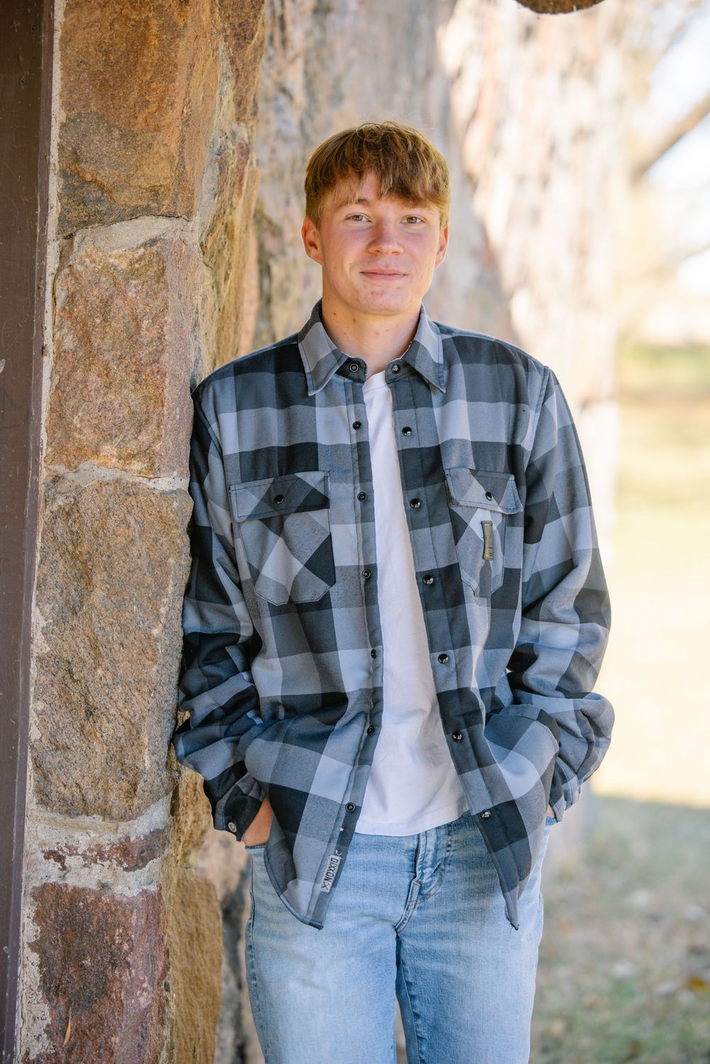 A teenage boy with red hair wearing a plaid flannel shirt and jeans leans against a stone wall outdoors.