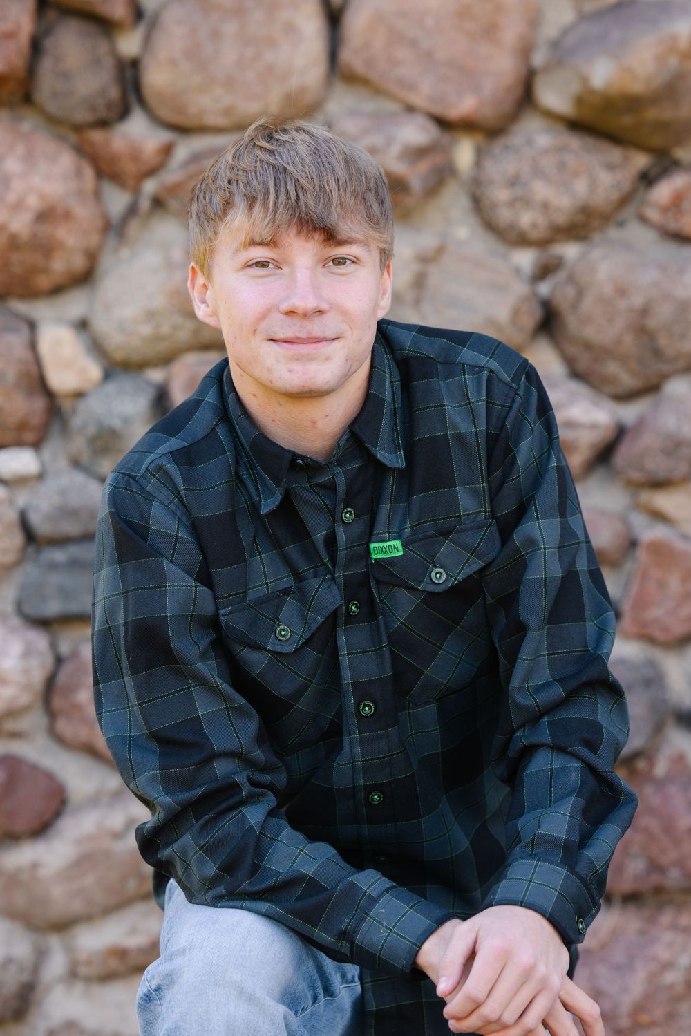 Young man in plaid flannel shirt smiling while sitting against a rustic stone wall outdoors.