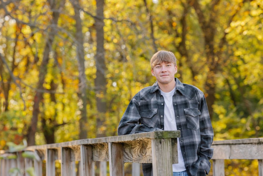 A teenage boy in a plaid flannel shirt leans on a wooden fence surrounded by vibrant yellow autumn foliage.