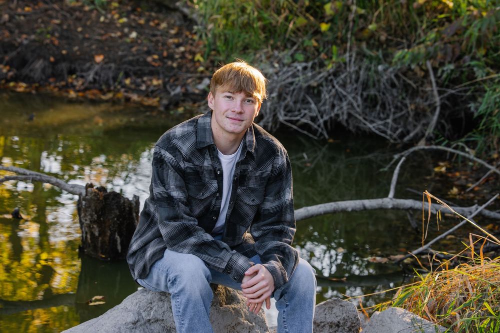 Young man with blonde hair sitting on rocks by a serene pond, wearing a plaid flannel shirt and jeans.