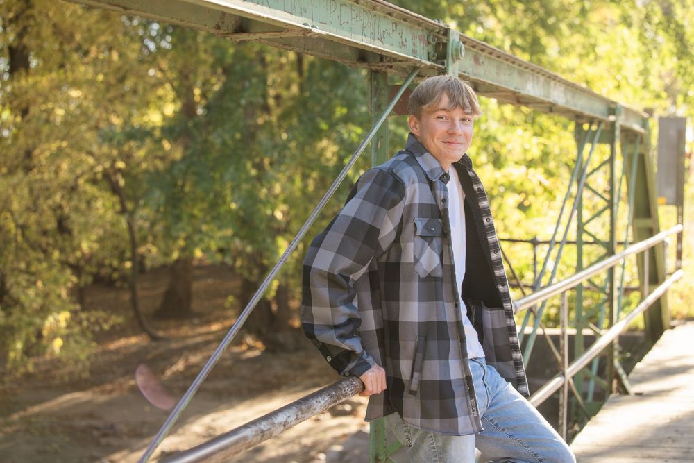Person in plaid shirt leaning on a green metal bridge railing surrounded by autumn trees.