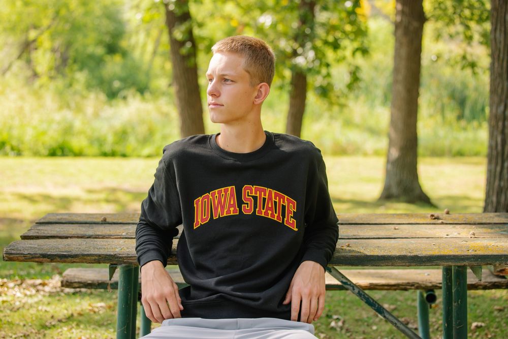 Young man wearing Iowa State sweatshirt sitting on a wooden bench outdoors surrounded by green trees.