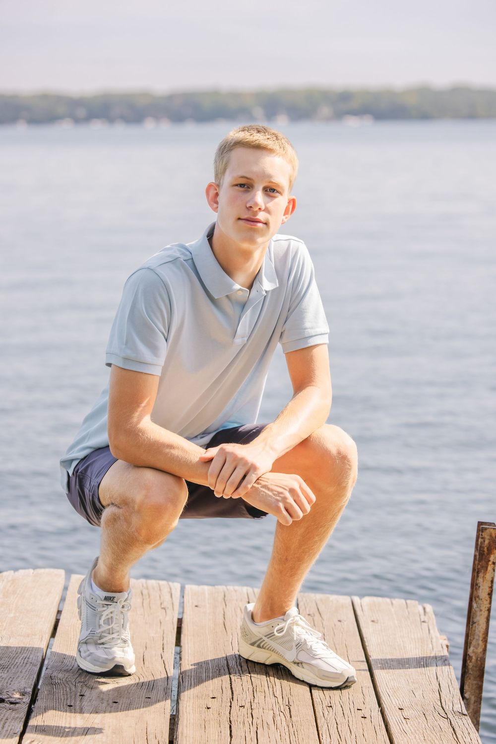 Young man in light blue polo and shorts crouching on a wooden dock with a calm lake in the background.
