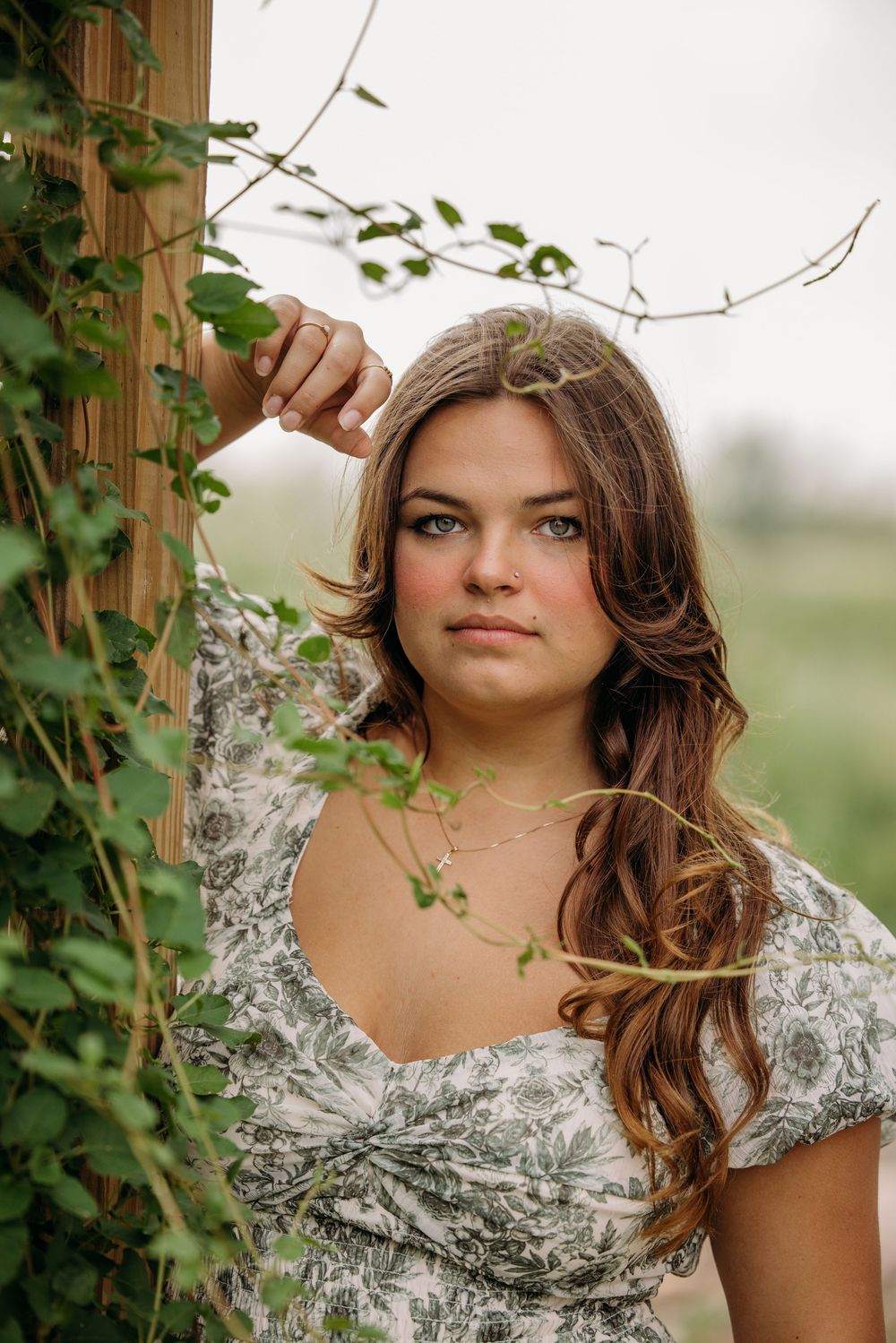 Young woman with long brown hair in floral dress leaning against wooden post surrounded by green vines outdoors.