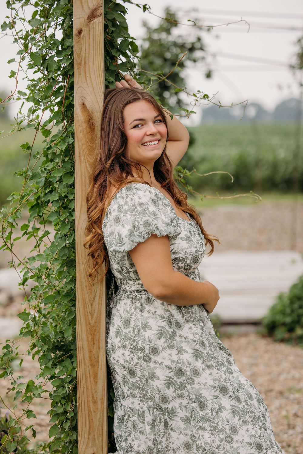 Young woman in floral dress leaning against wooden post with ivy, smiling outdoors in garden setting.
