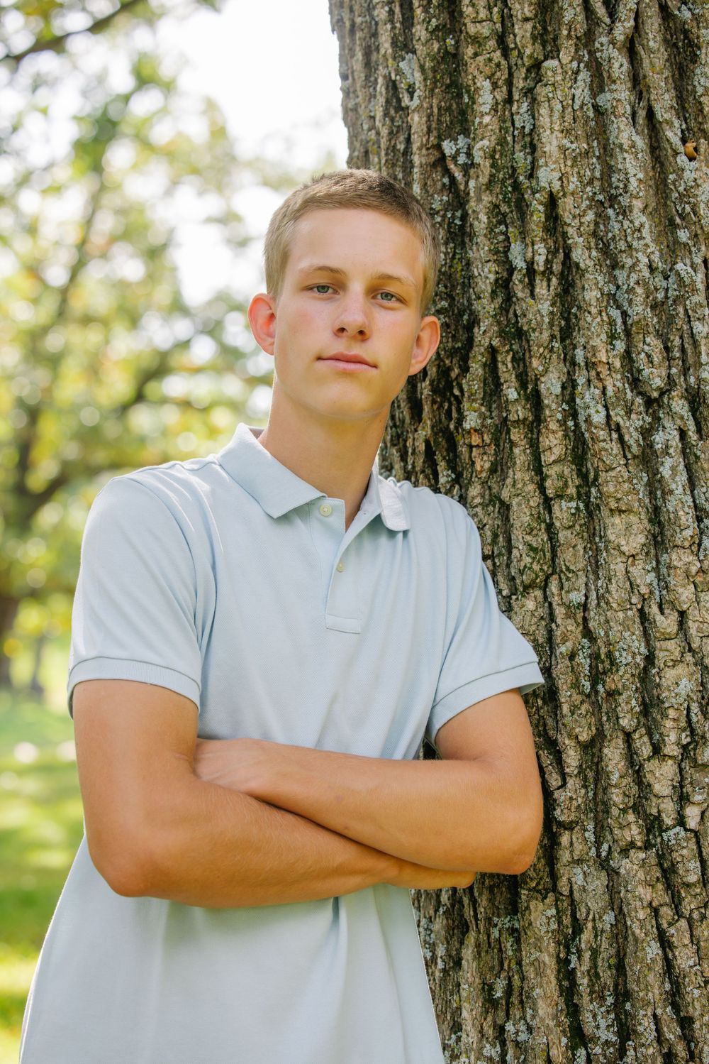 Young man in light blue polo shirt leaning against tree trunk with arms crossed in sunny park setting.