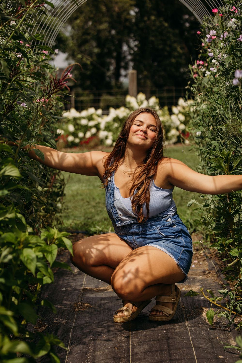 Young woman in denim overalls crouching with arms outstretched in a lush garden with blooming flowers.
