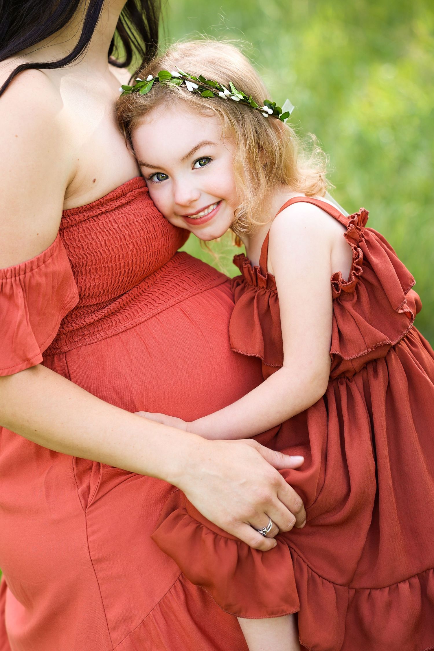 Smiling blonde girl in red dress with flower crown hugs her pregnant mothers belly outdoors.