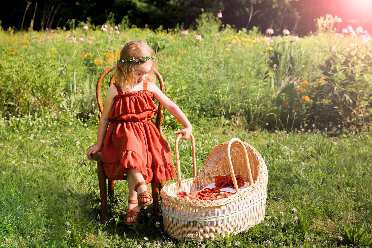 Toddler girl in red dress standing by wicker basket in sunny wildflower meadow.