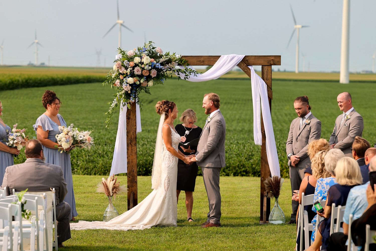 Outdoor wedding ceremony with couple at rustic wooden arch, wind turbines and green fields in background.