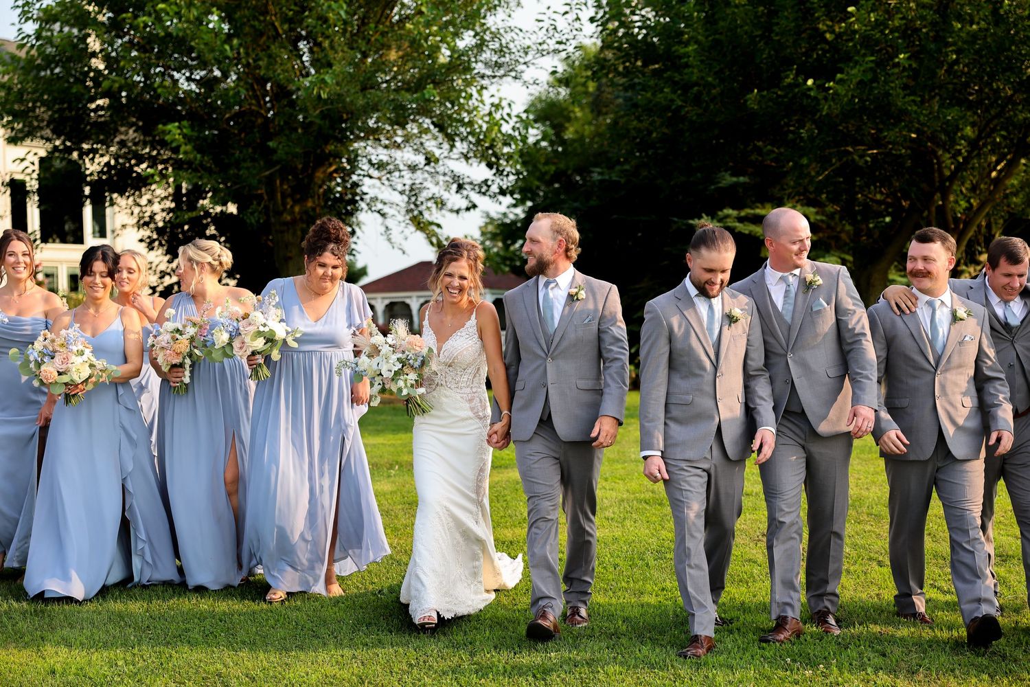 Wedding party walks together on green lawn, bridesmaids in blue gowns and groomsmen in gray suits with bride and groom.