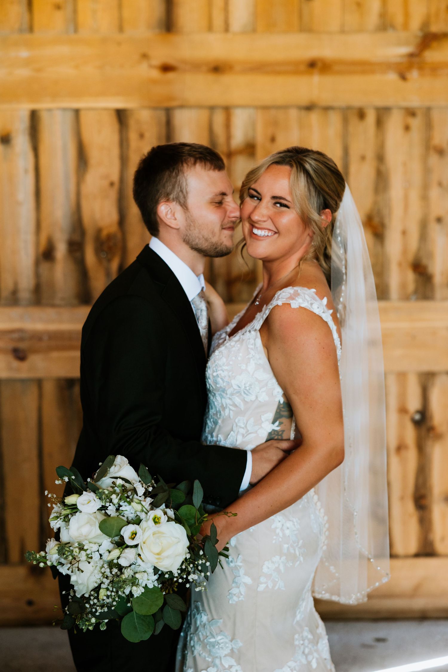 Bride smiles holding white floral bouquet as groom kisses her cheek inside a rustic wooden barn.