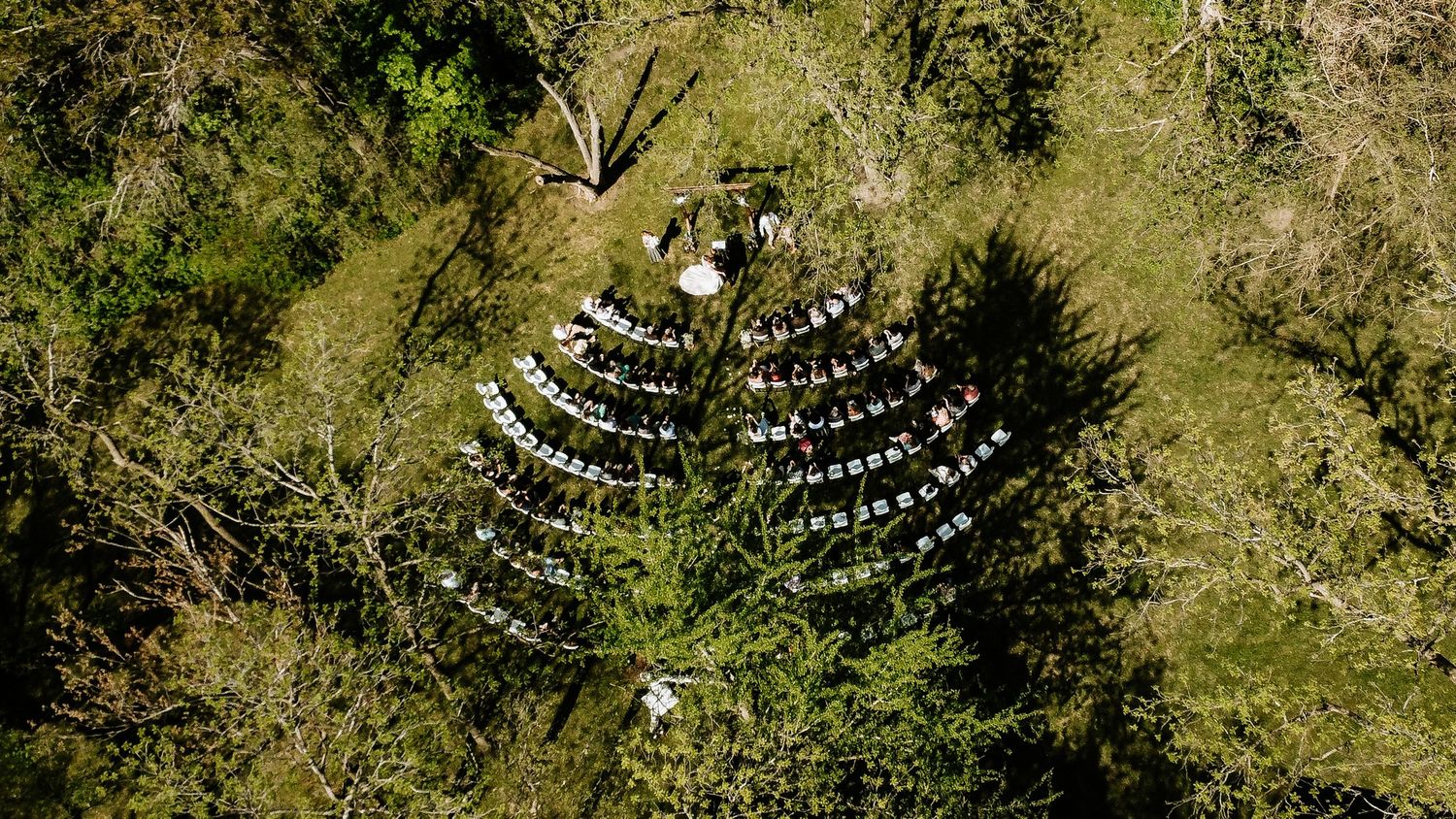 Aerial drone view of an outdoor wedding ceremony with white chairs arranged in a wooded forest clearing.