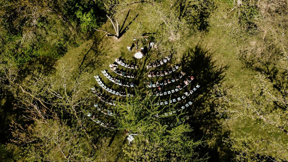 Aerial drone view of an outdoor wedding ceremony with white chairs arranged in a wooded forest clearing.