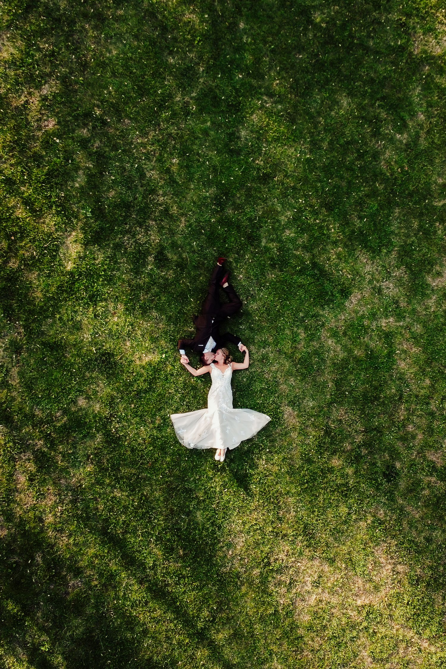 Aerial drone view of bride and groom lying on lush green grass in wedding attire.