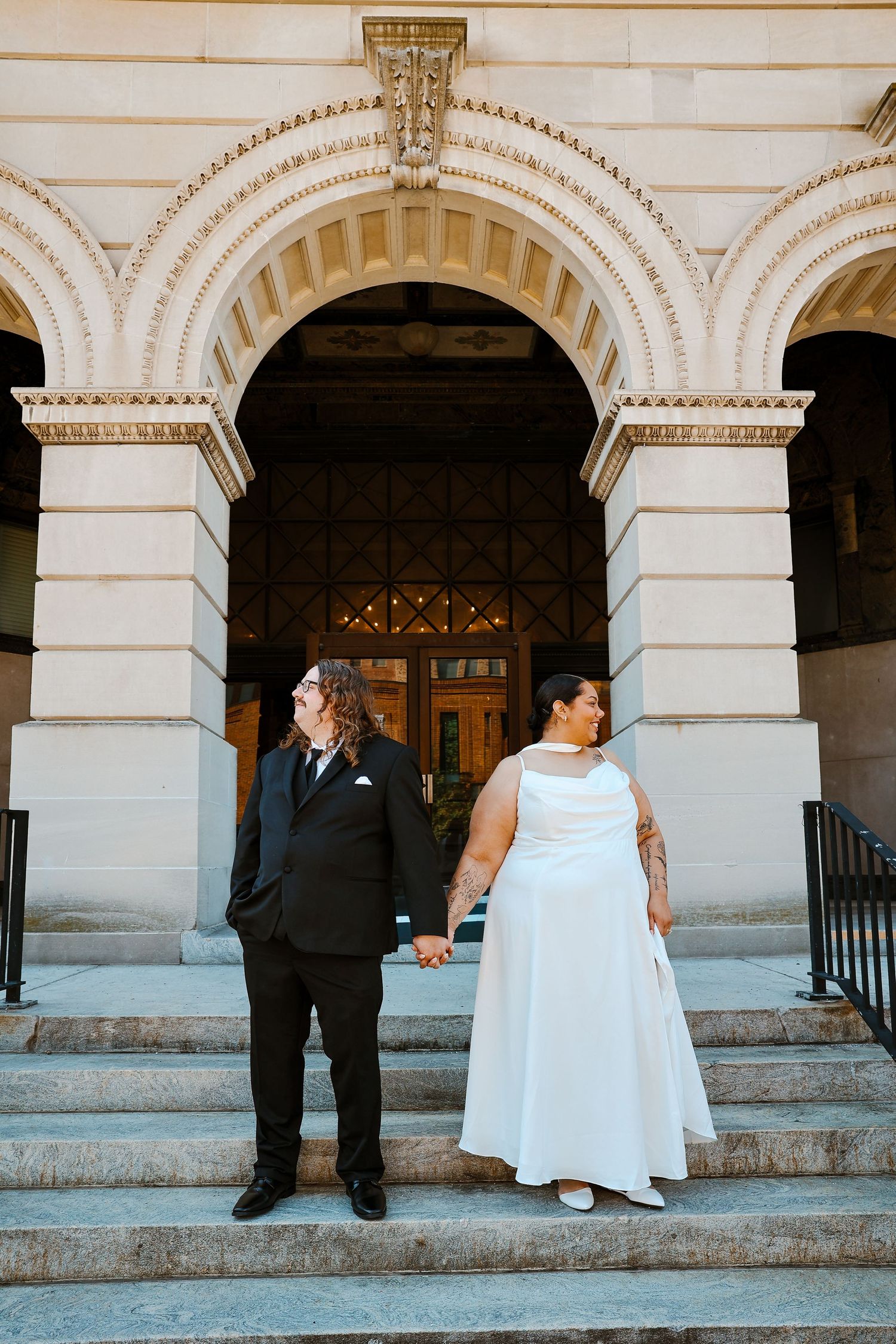 Bride and groom holding hands on steps of ornate stone building with arched entrance.