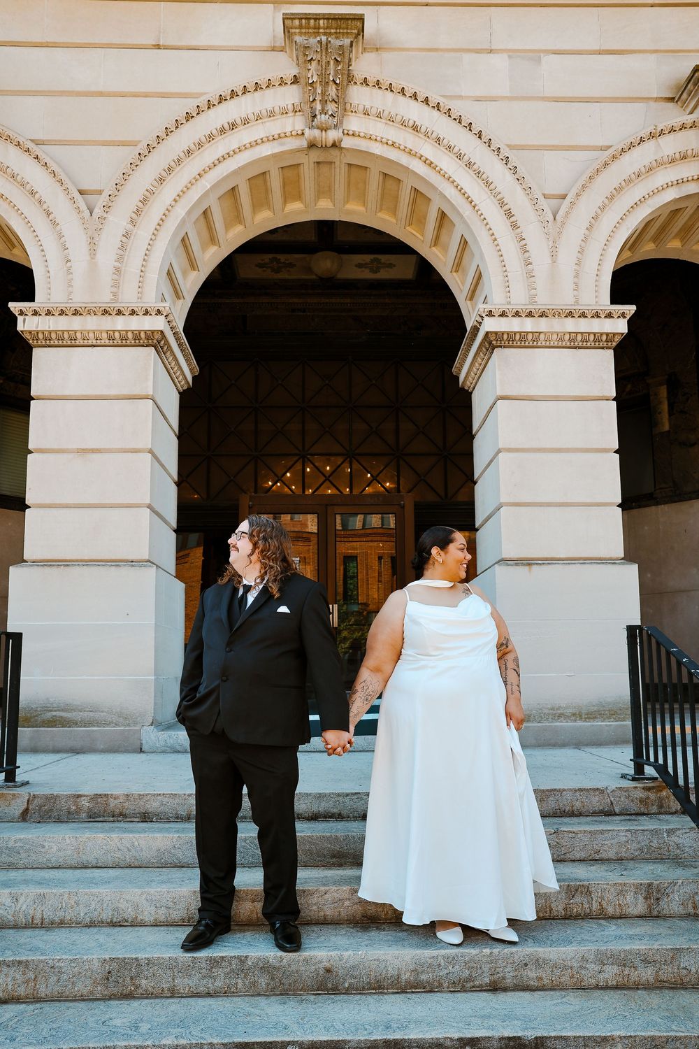 Bride and groom holding hands on steps of ornate stone building with arched entrance.