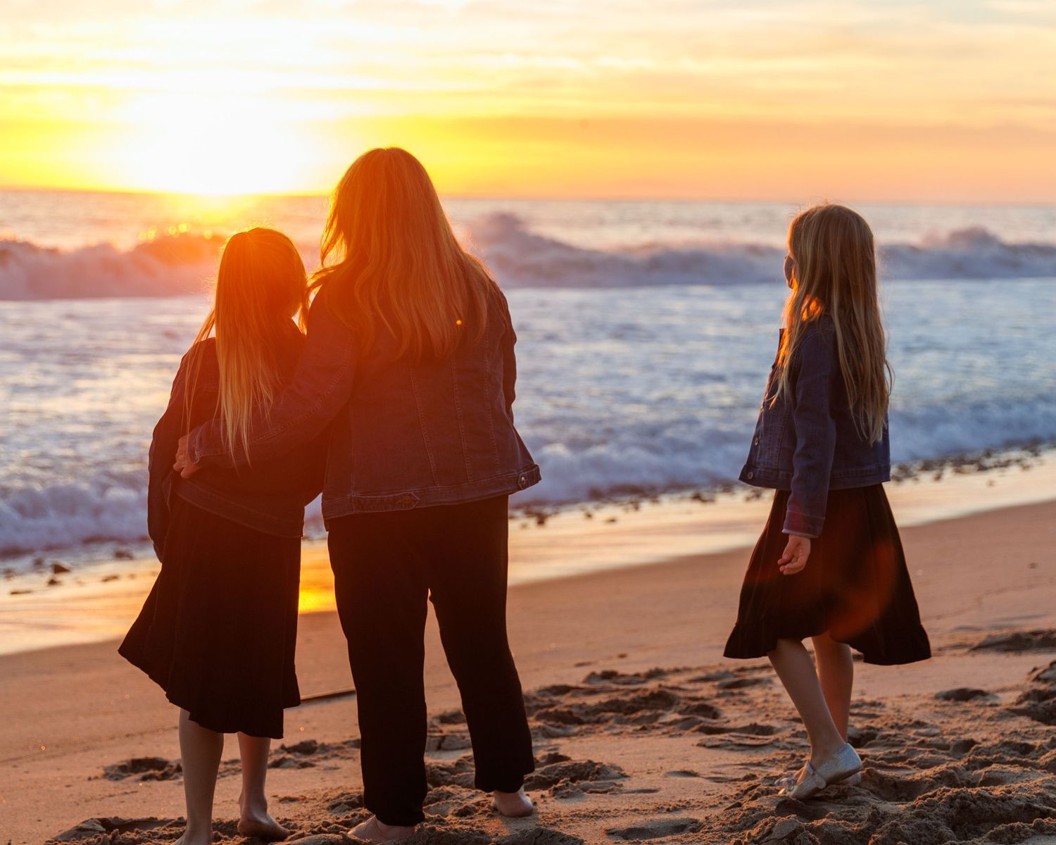 Candid family portrait of a mother and two daughters watch a stunning golden sunset over ocean waves on a sandy beach.
