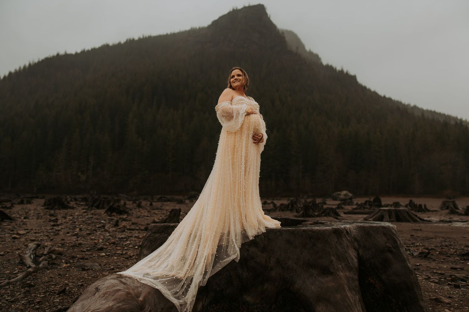 Pregnant woman in a flowing cream maternity gown stands on a rock before a dark misty mountain.