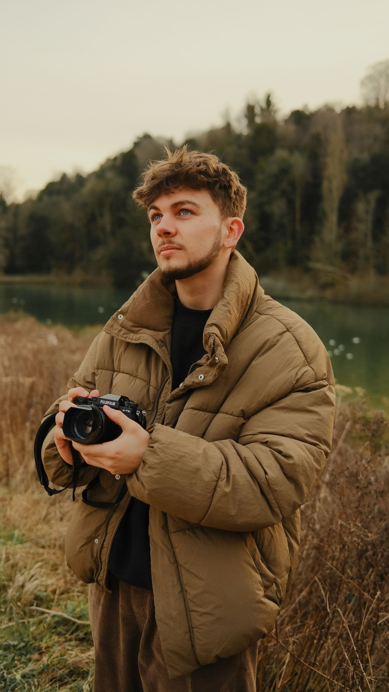A photographer in a brown winter coat holds his camera next to a lake while looking up