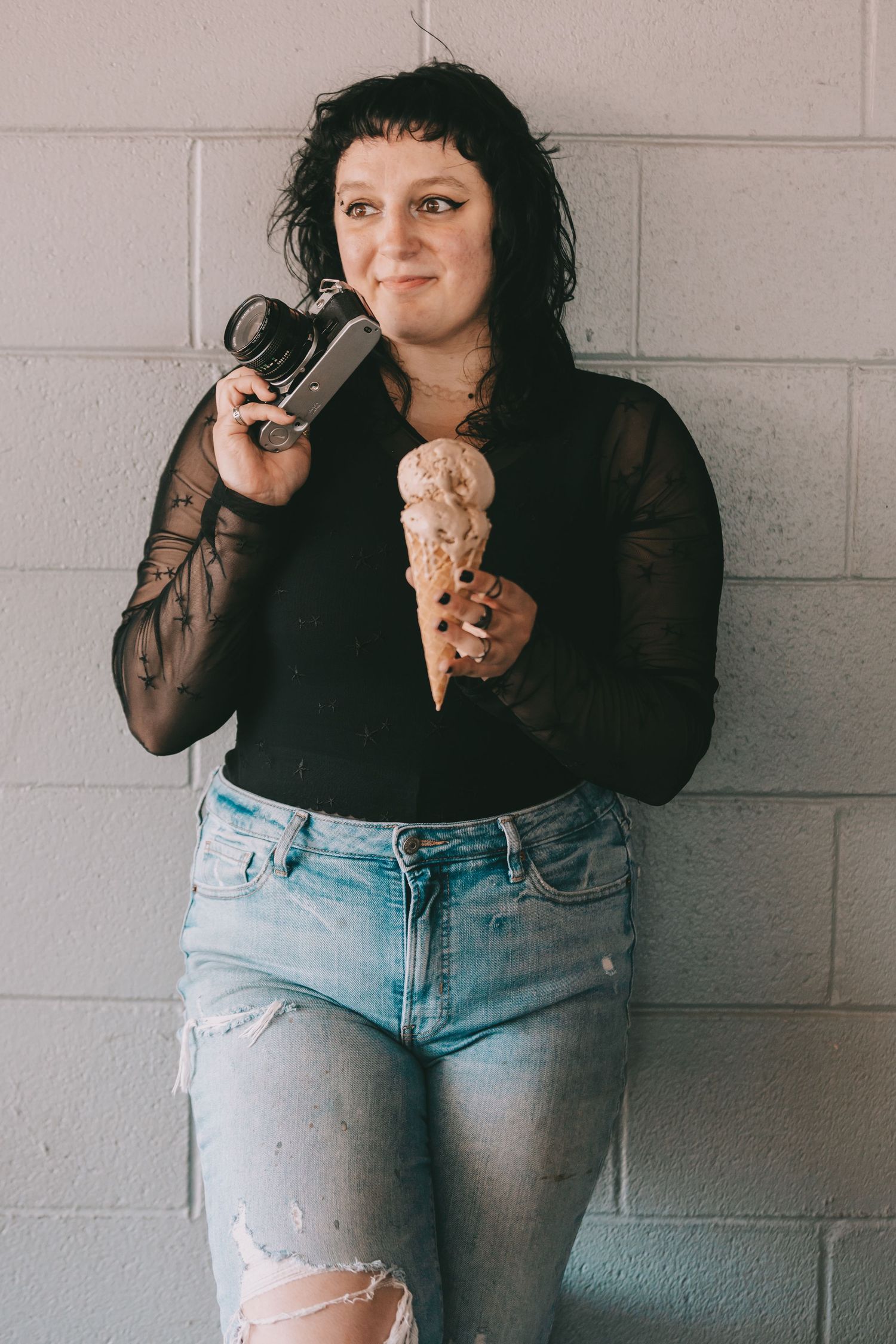 An image of a woman holding a camera and an ice cream cone standing in front of a grey brick wall.