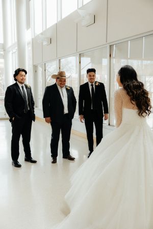 A wedding party in black suits and white dress pose in a bright modern space with windows.