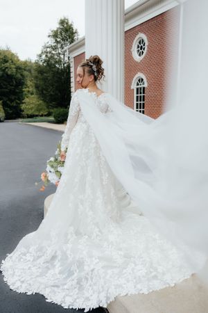 Stunning lace wedding dress with cathedral train flows against brick church backdrop.