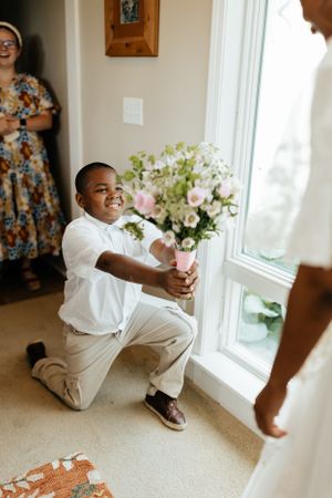 Young ring bearer in white shirt kneeling with flowers by window.
