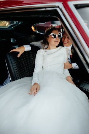 Bride in white dress and sunglasses sitting in vintage red car.