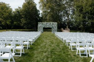 Outdoor wedding ceremony setup with white chairs and floral arch on a grassy lawn.