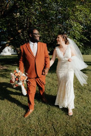 A couple in wedding attire walking together on grass, with the groom wearing a striking orange suit.