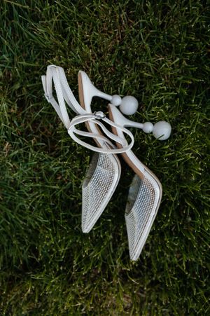 White lace bridal shoes with pearl details lying on grass.